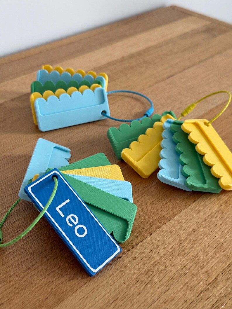 May include: A collection of rectangular plastic luggage tags in blue, green, and yellow, featuring scalloped edges and a smooth finish. One tag is prominently displayed with the name Leo printed in white text, attached to a green wire loop.