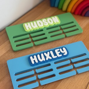 May include: Two rectangular wall-mounted medal hangers. The top hanger is green with the white name HUDSON on a light green background. The bottom hanger is blue with the white name HUXLEY on a dark blue background. Both feature horizontal slots.