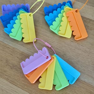 May include: Three sets of plastic yarn bobbins, each featuring a scalloped edge and a central hole for threading. The bobbins are arranged in fan-like clusters on flexible wire loops, showcasing a vibrant spectrum of colors including blue, green, yellow, orange, and purple.