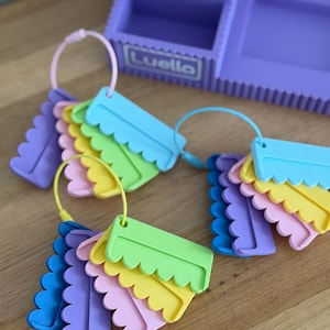 May include: A purple desk organizer with the name Luello printed on the front, accompanied by three circular rings holding sets of scalloped-edge plastic tags in various pastel colors including blue, green, yellow, pink, and purple.