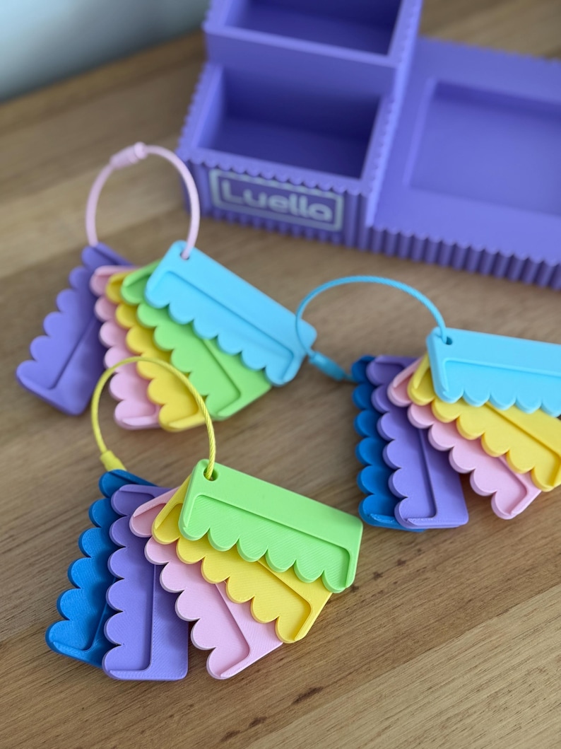 May include: A purple desk organizer with the name Luello printed on the front, accompanied by three circular rings holding sets of scalloped-edge plastic tags in various pastel colors including blue, green, yellow, pink, and purple.
