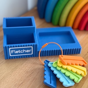 May include: A blue desk organizer with a rectangular base and two compartments, featuring the name Fletcher printed on the front. An orange ring holds five small, scalloped-edge plastic tags in blue, light blue, green, yellow, and orange.