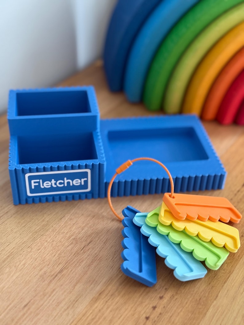 May include: A blue desk organizer with a rectangular base and two compartments, featuring the name Fletcher printed on the front. An orange ring holds five small, scalloped-edge plastic tags in blue, light blue, green, yellow, and orange.