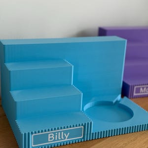 May include: A light blue tiered display stand featuring a three-step staircase on the left and a circular recessed holder on the right. The front of the bottom step displays a white rectangular label with the name Billy printed in black text.