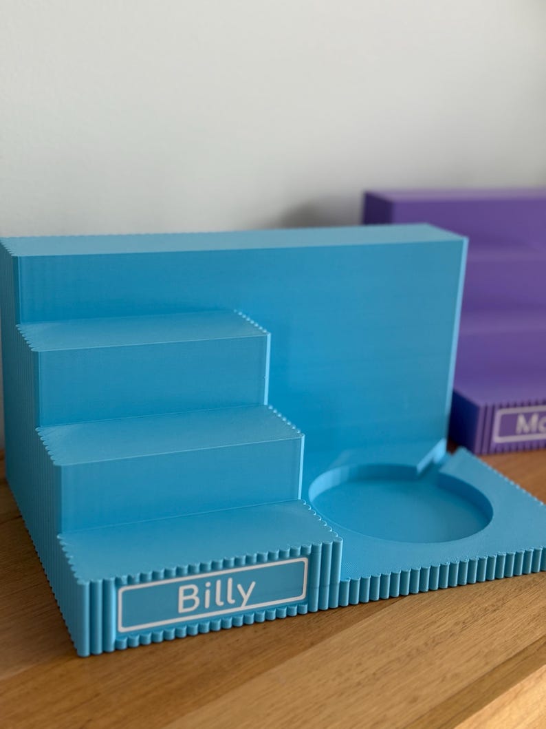 May include: A light blue tiered display stand featuring a three-step staircase on the left and a circular recessed holder on the right. The front of the bottom step displays a white rectangular label with the name Billy printed in black text.