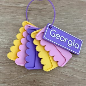 May include: A set of layered, scalloped-edge plastic tags in yellow, pink, and purple, held together by a purple wire loop. One rectangular purple tag features the name Georgia in white lettering, while the others are solid colored shapes.