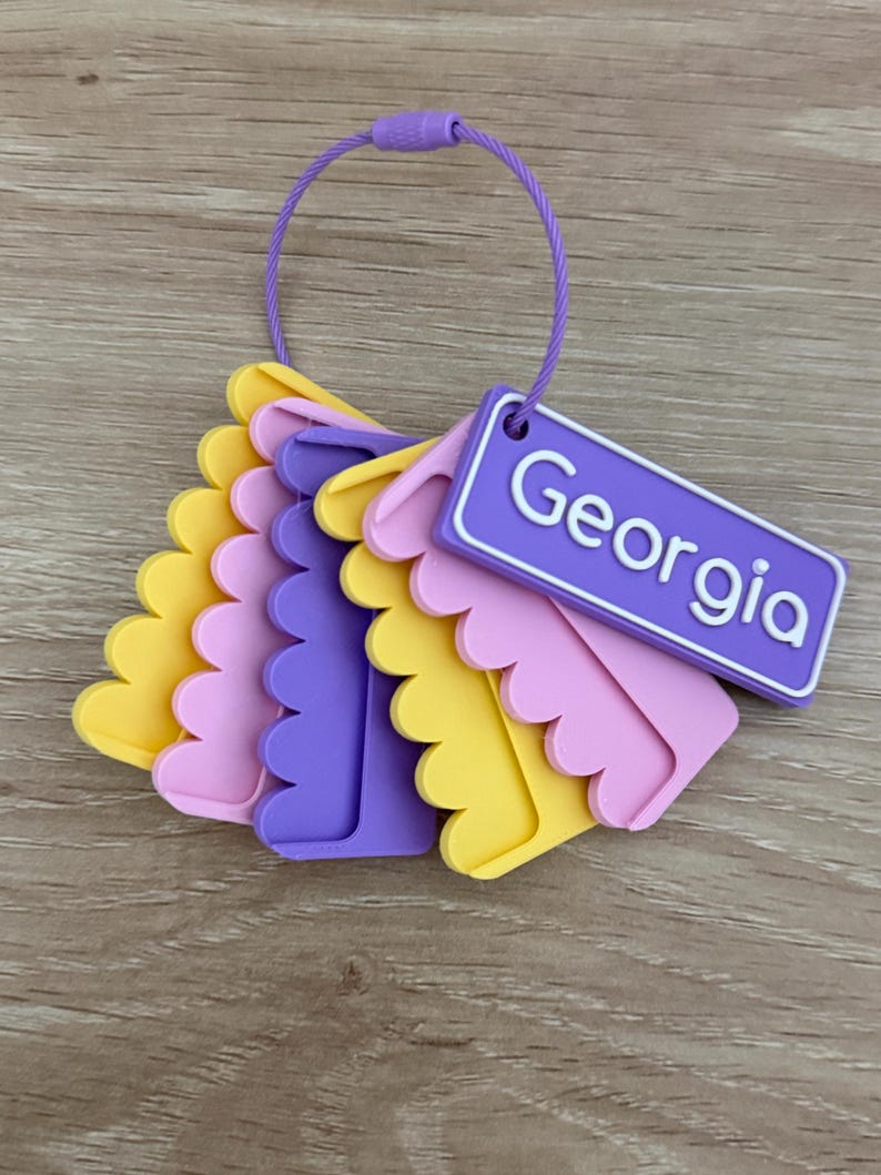 May include: A set of layered, scalloped-edge plastic tags in yellow, pink, and purple, held together by a purple wire loop. One rectangular purple tag features the name Georgia in white lettering, while the others are solid colored shapes.