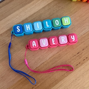 May include: Two personalised silicone bead keychains with white letters. One keychain spells SHILOH in blue and green beads with a blue cord. The other spells AVERY in pink and light pink beads with a matching pink cord attached to the side.