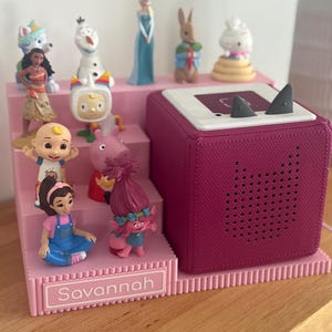 May include: A pink tiered display stand with the name Savannah printed on the front, holding various small character figurines. Next to the stand is a square magenta speaker with a white top surface featuring two small grey triangular ears.
