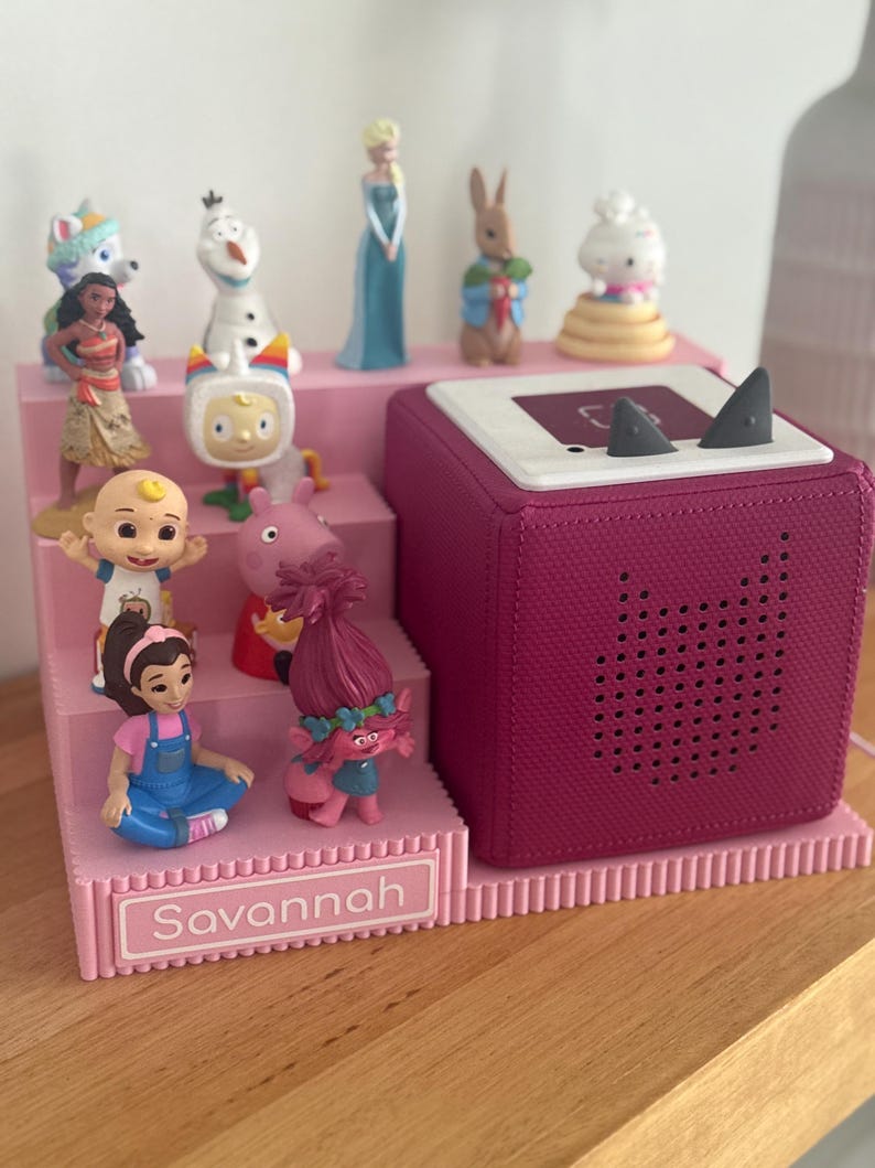 May include: A pink tiered display stand with the name Savannah printed on the front, holding various small character figurines. Next to the stand is a square magenta speaker with a white top surface featuring two small grey triangular ears.