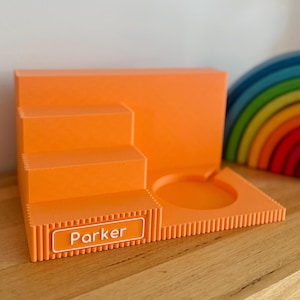 May include: A bright orange tiered display stand featuring a stepped design on the left and a circular cutout on the right. The front edge has a rectangular white nameplate with the name Parker printed in white text against an orange background.