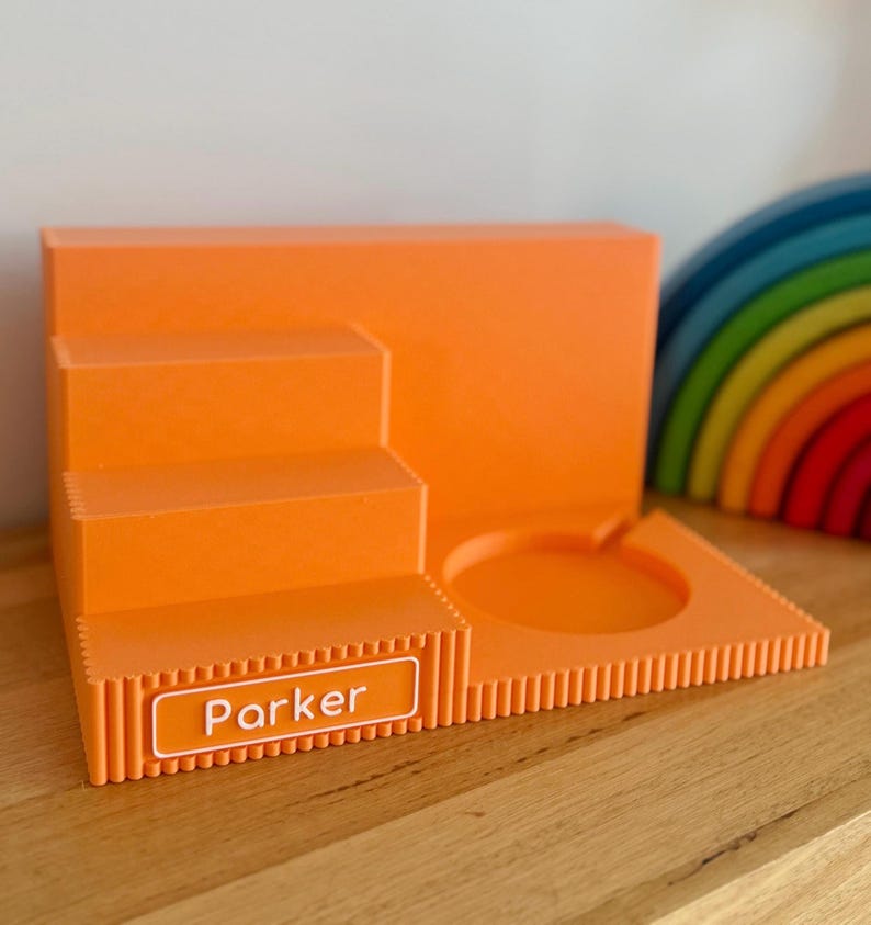 May include: A bright orange tiered display stand featuring a stepped design on the left and a circular cutout on the right. The front edge has a rectangular white nameplate with the name Parker printed in white text against an orange background.
