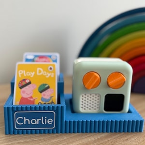 May include: A blue plastic storage base with the name Charlie printed on the front, holding two rectangular cards featuring Peppa Pig characters and the text Play Days, alongside a light green audio player with two orange knobs and a speaker.