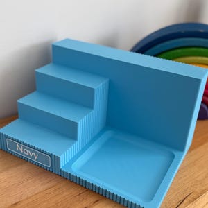 May include: A light blue tiered display stand with a stepped design and a flat square base. The front edge features a rectangular label with the text Navy printed in white. The structure has a ribbed texture along the sides and edges.