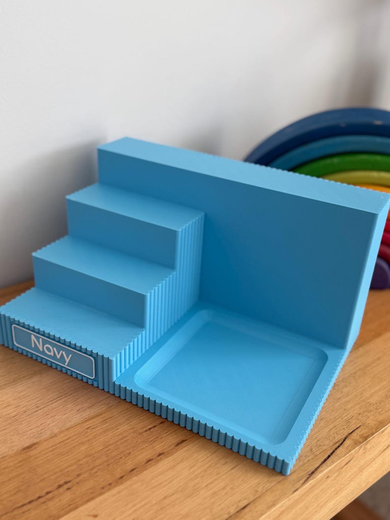 May include: A light blue tiered display stand with a stepped design and a flat square base. The front edge features a rectangular label with the text Navy printed in white. The structure has a ribbed texture along the sides and edges.