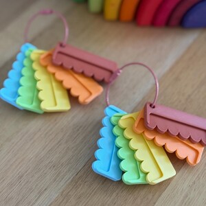 May include: Two sets of colorful, scalloped-edge rectangular tags arranged on thin pink metal rings. Each set includes blue, green, yellow, orange, and muted red pieces. The tags feature a distinct wavy bottom edge and a smooth, flat surface.