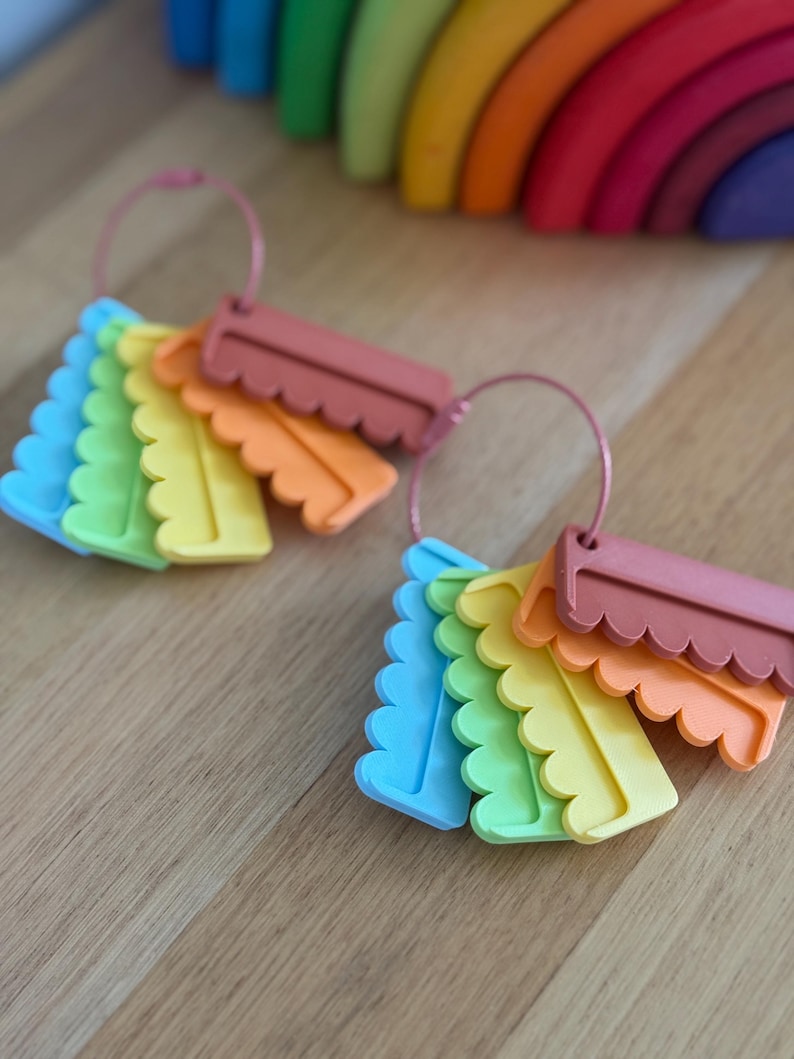 May include: Two sets of colorful, scalloped-edge rectangular tags arranged on thin pink metal rings. Each set includes blue, green, yellow, orange, and muted red pieces. The tags feature a distinct wavy bottom edge and a smooth, flat surface.