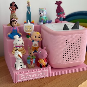 May include: A pink tiered display stand with the name Savannah printed on the front, holding a collection of small character figurines and a pink square speaker with two small grey ears on top and a perforated cat-shaped design on the front.