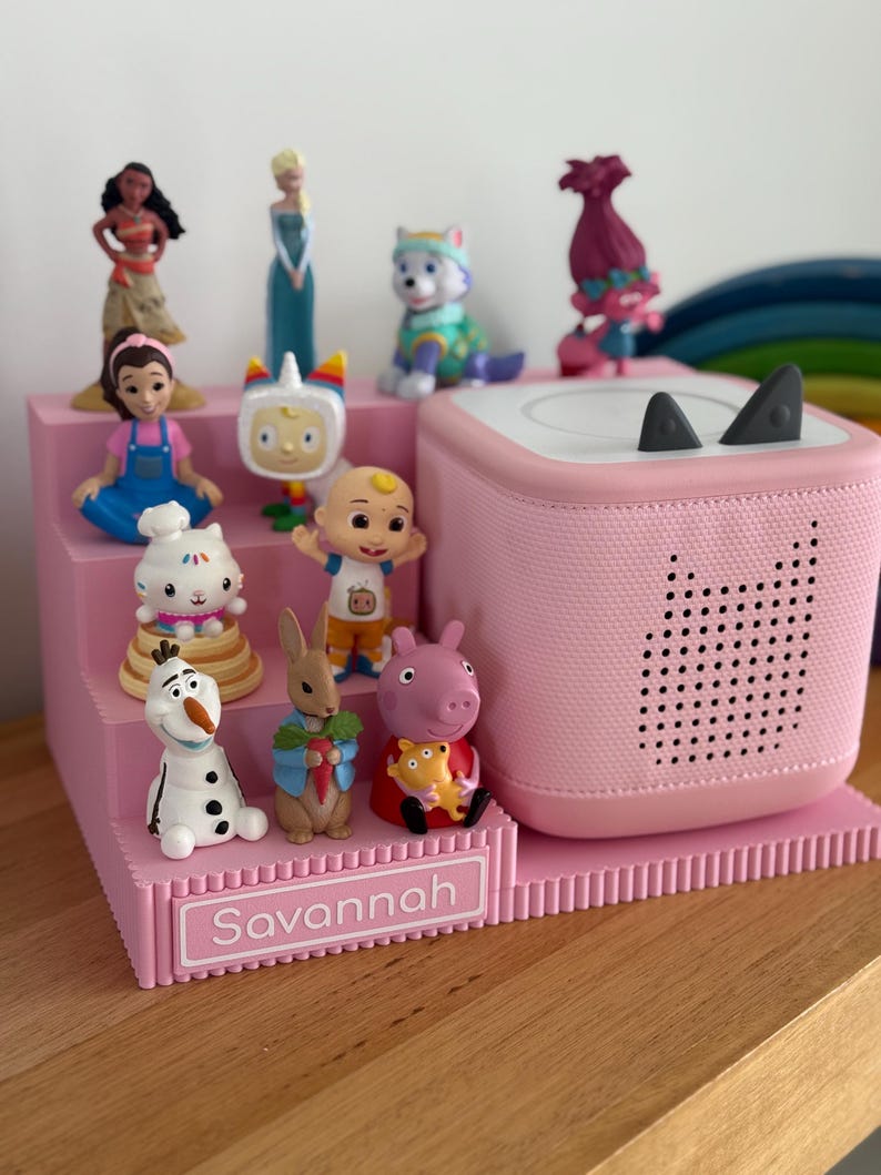 May include: A pink tiered display stand with the name Savannah printed on the front, holding a collection of small character figurines and a pink square speaker with two small grey ears on top and a perforated cat-shaped design on the front.