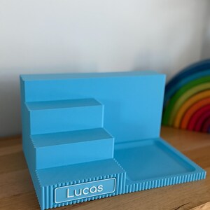 May include: A light blue tiered display stand featuring a stepped design with three levels and a flat square base. The front edge of the lowest step displays a rectangular nameplate with the white text Lucas printed in a clean sans-serif font.