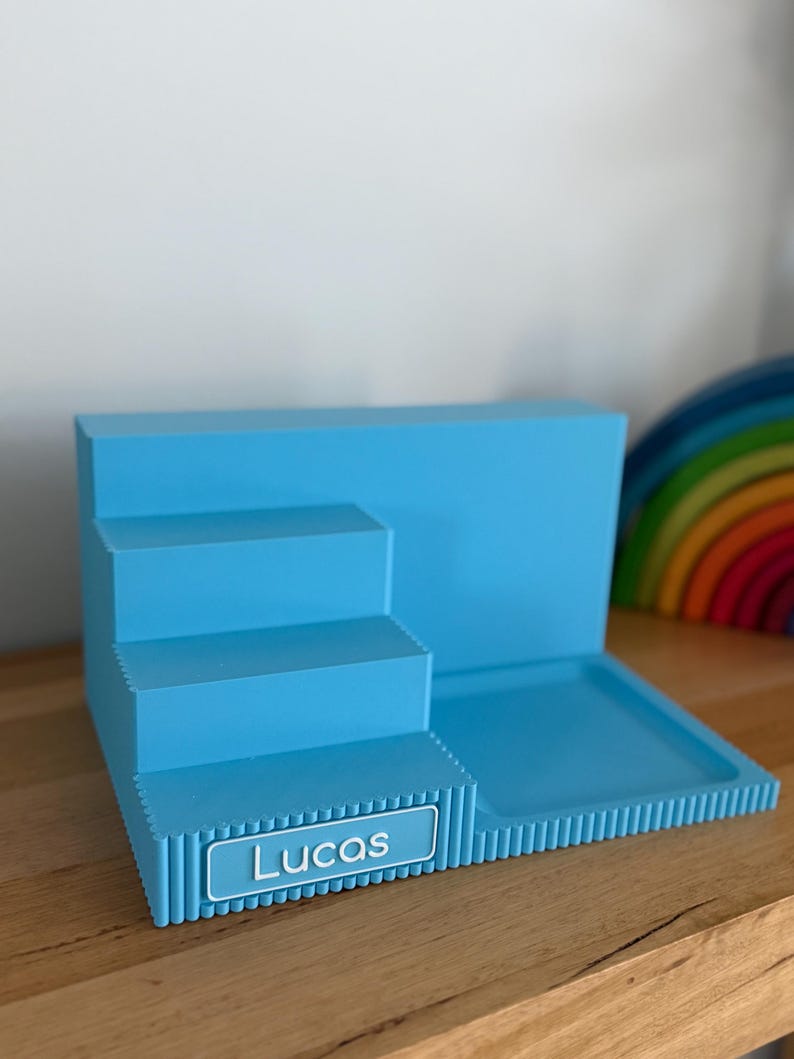 May include: A light blue tiered display stand featuring a stepped design with three levels and a flat square base. The front edge of the lowest step displays a rectangular nameplate with the white text Lucas printed in a clean sans-serif font.