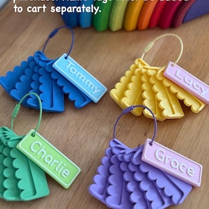 May include: Four sets of scalloped plastic card clips in blue, yellow, green, and purple, each attached to a matching wire ring. Rectangular name tags labeled Tommy, Lucy, Charlie, and Grace are attached. Text reads: *Name tags not included in card clip purchase. Name tags must be added to cart separately.