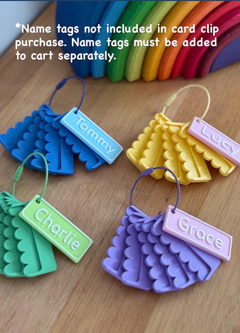 May include: Four sets of scalloped plastic card clips in blue, yellow, green, and purple, each attached to a matching wire ring. Rectangular name tags labeled Tommy, Lucy, Charlie, and Grace are attached. Text reads: *Name tags not included in card clip purchase. Name tags must be added to cart separately.