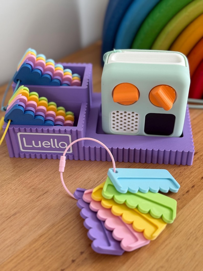 May include: A purple plastic toy organizer with the name Luella printed on the front. It holds two sets of rainbow-colored scalloped blocks and a mint green square music player with two orange knobs, a speaker grid, and a black screen. A ring of pastel tags.