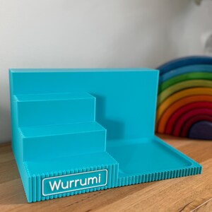 May include: A bright turquoise plastic display stand featuring a three-tiered staircase design on the left and a flat rectangular platform on the right. The front edge of the bottom step displays a white rectangular label with the text Wurrumi.