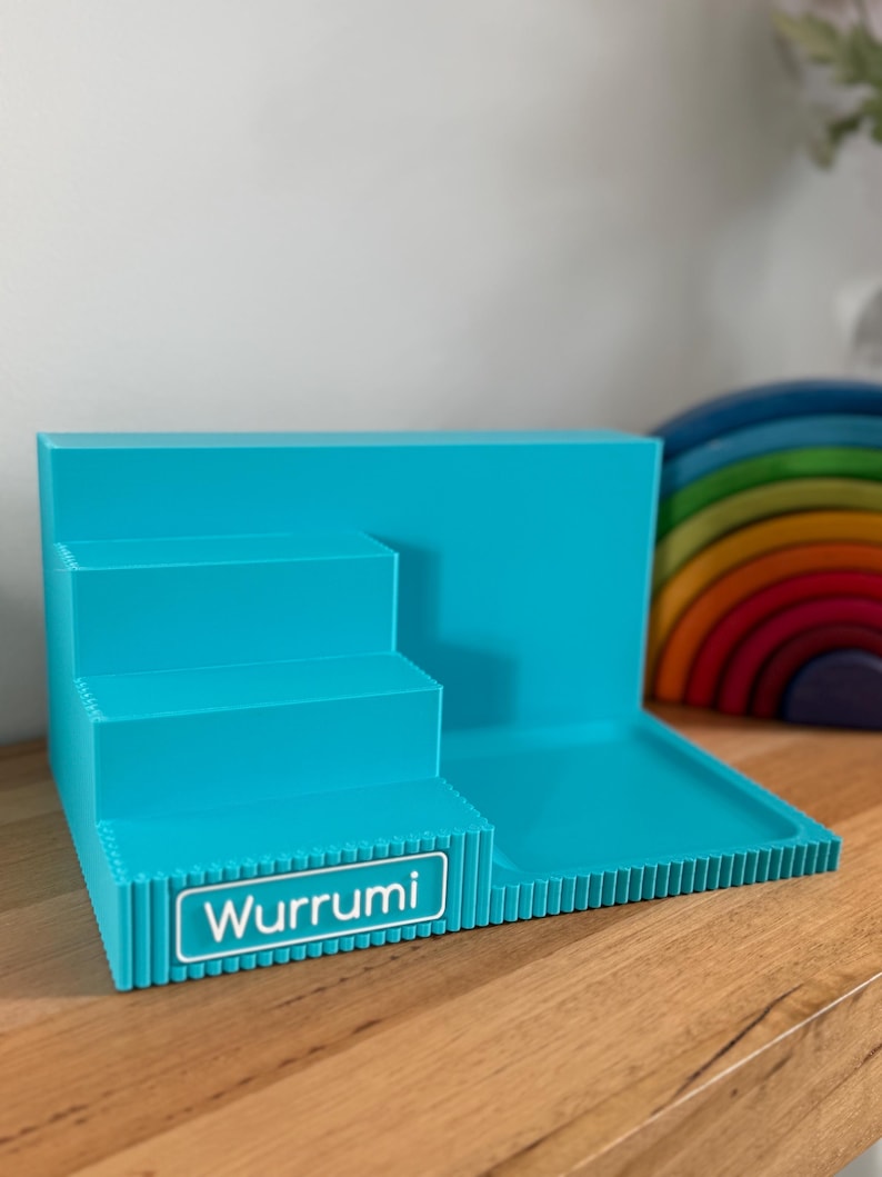 May include: A bright turquoise plastic display stand featuring a three-tiered staircase design on the left and a flat rectangular platform on the right. The front edge of the bottom step displays a white rectangular label with the text Wurrumi.