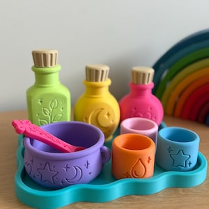 May include: A set of colourful silicone potion-making toys including a light green bottle, a yellow bottle with a moon, a pink bottle, a purple cauldron with a matching spoon, and three small cups in orange, pink, and blue, all on a teal cloud-shaped tray.