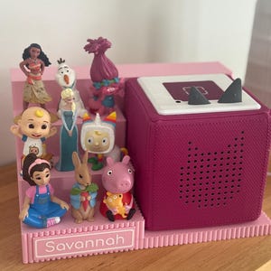 May include: A pink tiered display stand with the name Savannah printed on the front, holding various small character figurines. Next to the stand is a dark pink square audio speaker with a white top featuring two small grey triangular ears.