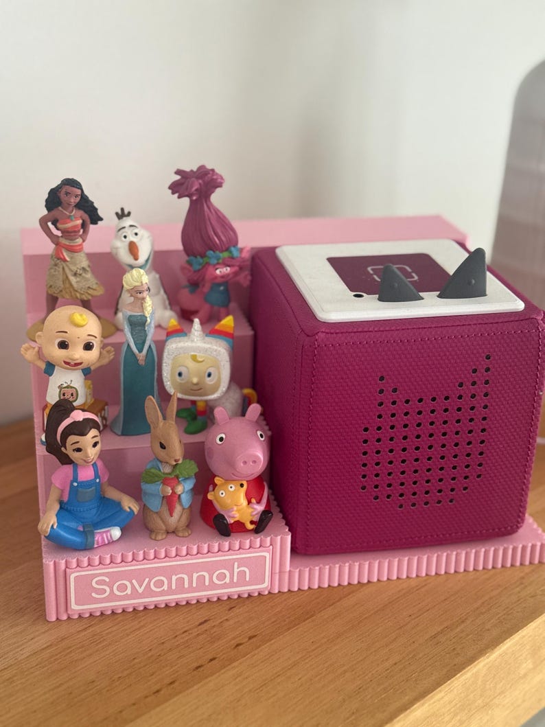May include: A pink tiered display stand with the name Savannah printed on the front, holding various small character figurines. Next to the stand is a dark pink square audio speaker with a white top featuring two small grey triangular ears.