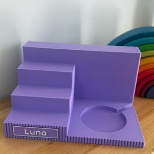 May include: A lavender-coloured tiered display stand featuring a staircase design on the left and a circular cutout on the right. The front edge of the bottom step displays a rectangular label with the name Luna printed in white text.