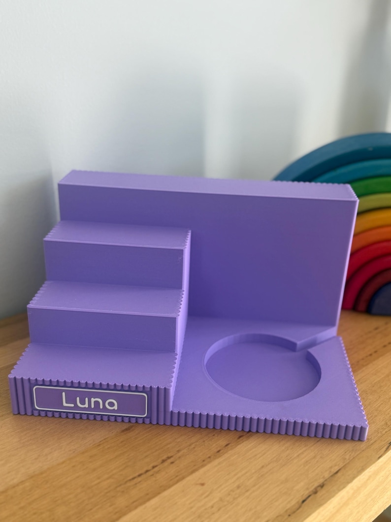 May include: A lavender-coloured tiered display stand featuring a staircase design on the left and a circular cutout on the right. The front edge of the bottom step displays a rectangular label with the name Luna printed in white text.