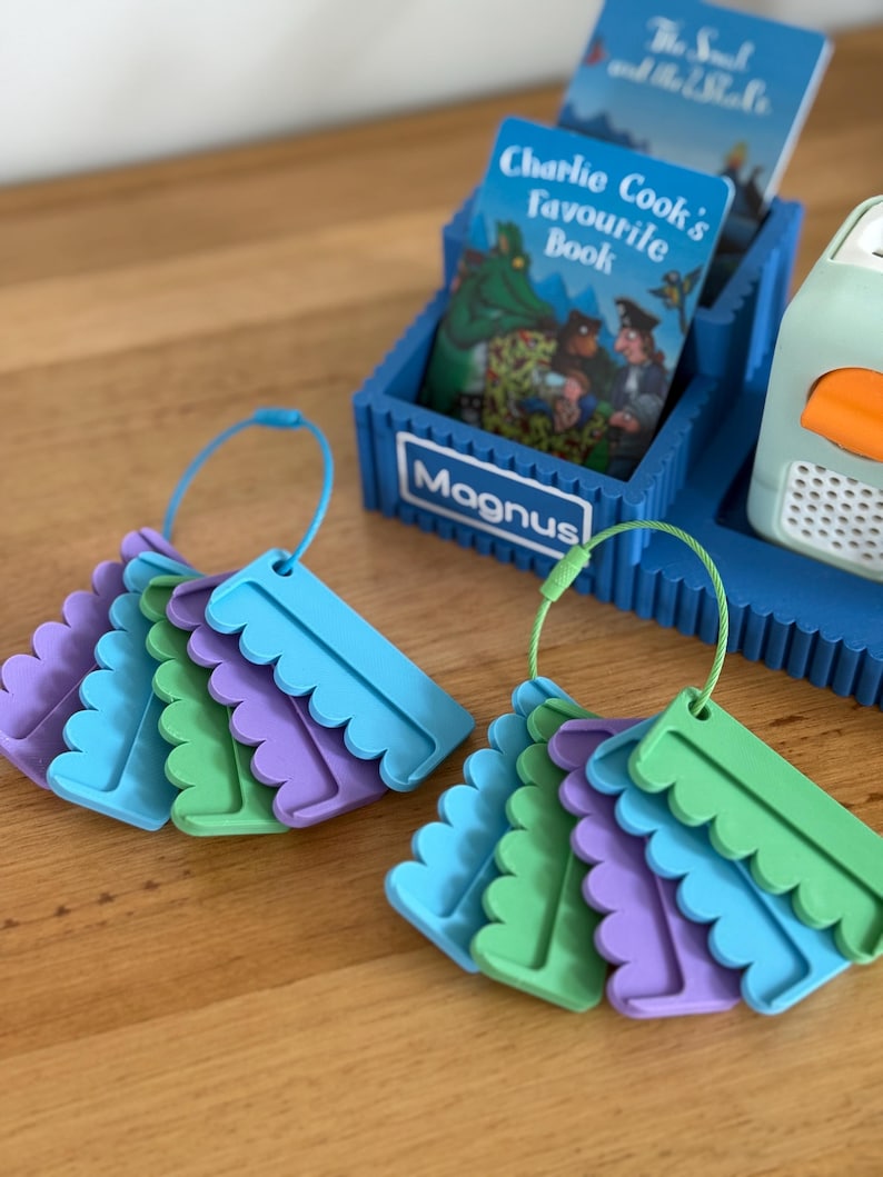 May include: Two sets of colorful plastic tags in blue, green, and purple, each attached to a circular wire ring. A blue plastic storage stand with the name Magnus printed on the front holds a book titled Charlie Cook's Favourite Book.