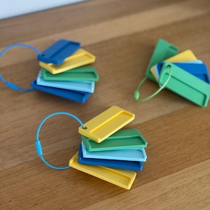 May include: Three sets of rectangular plastic tags in shades of blue, green, and yellow, each held together by a flexible metal cable ring. The tags feature a raised border and a small hole for the cable, arranged in a fanned-out, layered display.