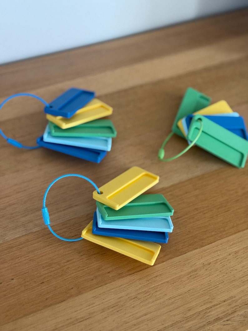 May include: Three sets of rectangular plastic tags in shades of blue, green, and yellow, each held together by a flexible metal cable ring. The tags feature a raised border and a small hole for the cable, arranged in a fanned-out, layered display.