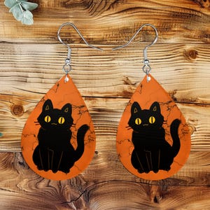 May include: A pair of teardrop-shaped earrings featuring a black cat illustration with yellow eyes on an orange background. The earrings have silver-colored hooks and are designed for pierced ears. The cat design is a Halloween theme.