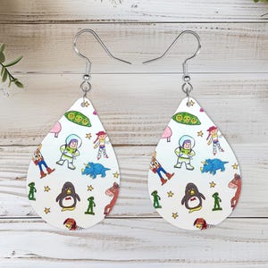 May include: A pair of teardrop-shaped earrings featuring a white background with a colorful repeating pattern of cartoon characters including a space ranger, a cowgirl, a penguin, a dinosaur, a horse, and green peas, finished with silver-toned hooks.