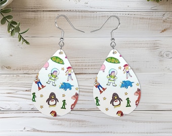 May include: A pair of teardrop-shaped earrings featuring a white background with a colorful repeating pattern of cartoon characters including a space ranger, a cowgirl, a penguin, a dinosaur, a horse, and green peas, finished with silver-toned hooks.