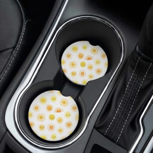 May include: Two circular car cup holder coasters featuring a repeating pattern of yellow and orange suns with simple, smiling faces on a white background. The coasters are positioned inside the black plastic cup holders of a vehicle console.