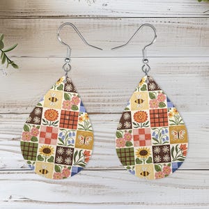 May include: A pair of teardrop-shaped dangle earrings featuring a patchwork pattern of squares. The design includes illustrations of sunflowers, daisies, bluebells, pink flowers, bees, and butterflies in shades of yellow, orange, green, and blue.