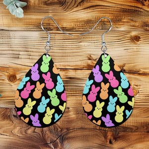 May include: A pair of teardrop-shaped earrings with a black background and a pattern of colorful Easter bunny shapes. The bunnies are in shades of pink, blue, green, yellow, orange, and purple. The earrings have silver-colored hooks.