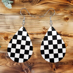 May include: A pair of teardrop-shaped earrings with a black and white checkered pattern. The earrings are suspended from silver-colored fish hook ear wires. The earrings are displayed on a wooden surface.