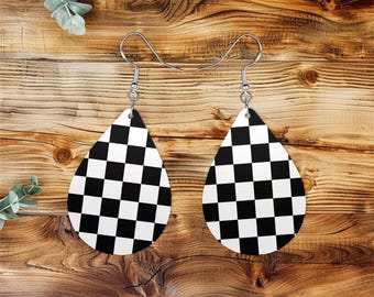 May include: A pair of teardrop-shaped earrings with a black and white checkered pattern. The earrings are suspended from silver-colored fish hook ear wires. The earrings are displayed on a wooden surface.