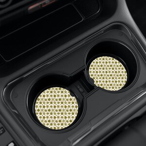 May include: Two circular car cup holder coasters featuring a repeating pattern of small, olive green flowers and dots on a cream-colored background. The coasters are positioned inside the black plastic cup holders of a vehicle center console.