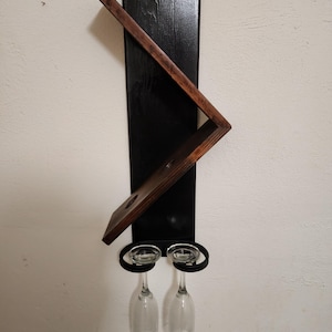 May include: A wall-mounted wine rack with a black wooden backboard and a dark brown angled shelf. Two clear wine glasses hang below in black metal holders. The rack is designed to hold a wine bottle and two glasses.