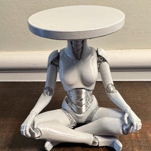 May include: A white and silver figurine of a seated humanoid robot with a round tabletop head. The robot is in a cross-legged position, with articulated joints and a detailed metallic torso. The tabletop is painted white.
