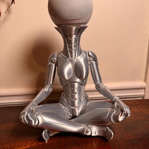 May include: A silver-colored, 3D-printed robotic figure in a seated, cross-legged position. The figure features a smooth, metallic finish with segmented armor plating and a hollow neck area designed to hold a spherical, light gray smart speaker.